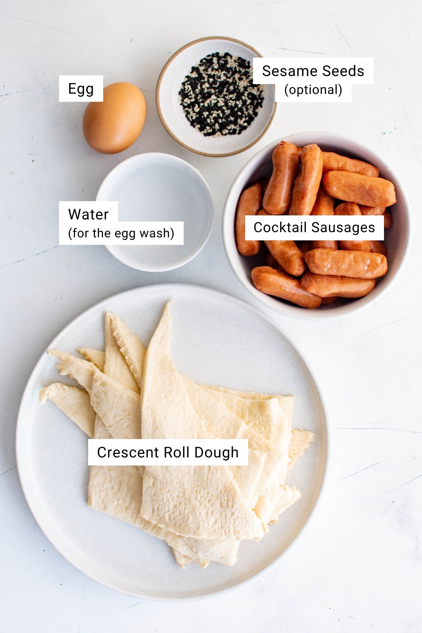 Ingredients for pigs in a blanket including crescent roll dough, cocktail sausages, egg, water, and sesame seeds