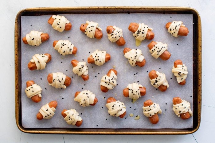 Pigs in a blanket topped with sesame seeds before baking on a parchment-lined baking sheet