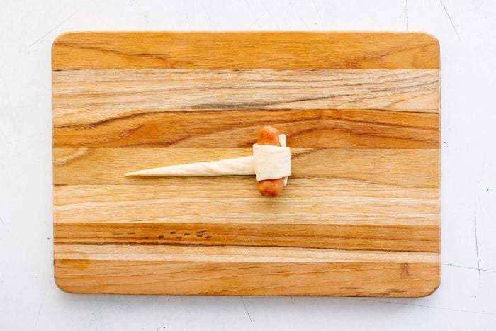 A cocktail sausage partially wrapped in crescent roll dough, showing how to start rolling pigs in a blanket.