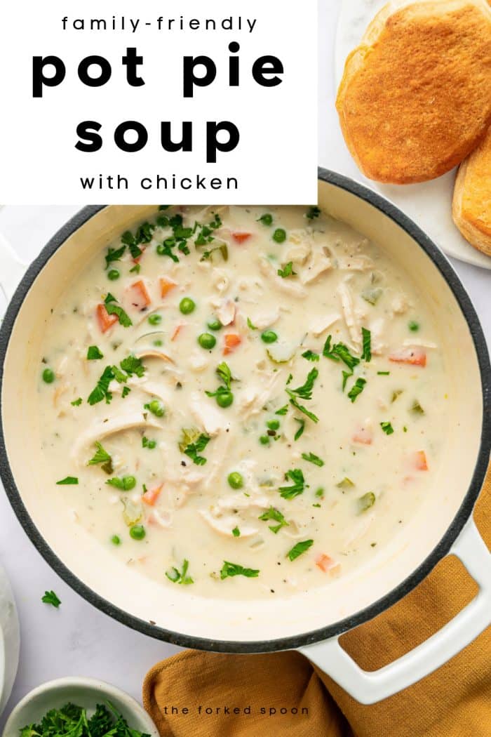 Creamy chicken pot pie soup in a white pot with vegetables and parsley, served with golden biscuits on the side.