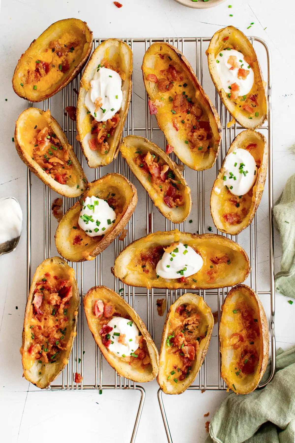 Thirteen potato skins filled with cheese, bacon, chives, and some with sour cream on a wire cooking rack.