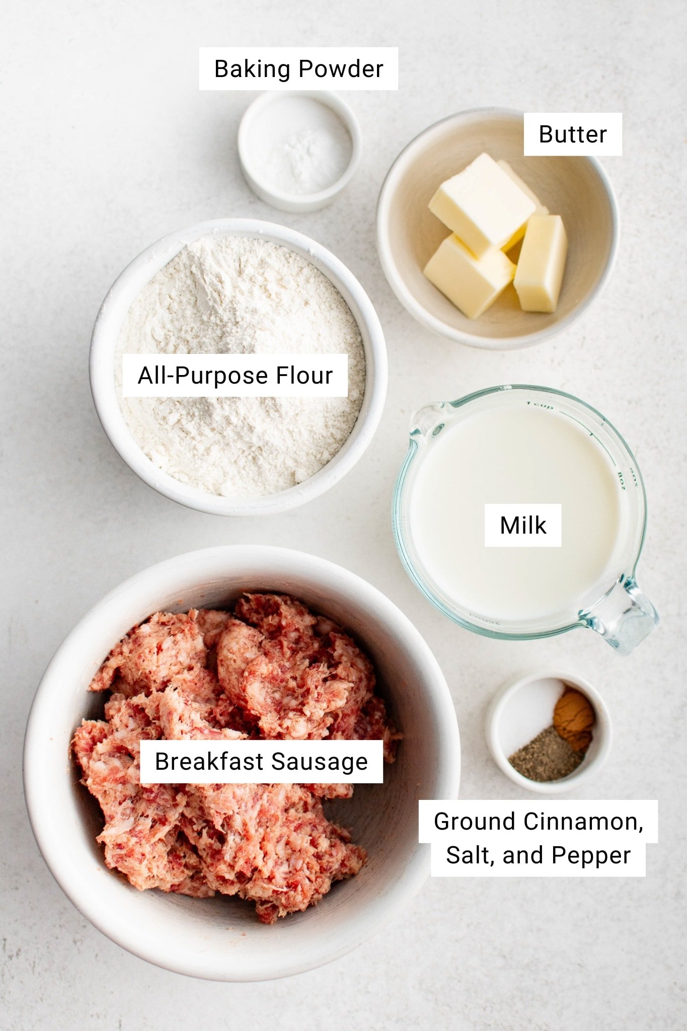 Overhead view of labeled ingredients for biscuits and gravy including flour, baking powder, butter, milk, breakfast sausage, and spices.