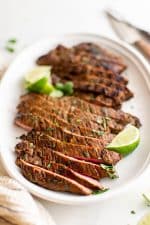 Large serving plate filled with thinly sliced carne asada garnished with limes and fresh cilantro.