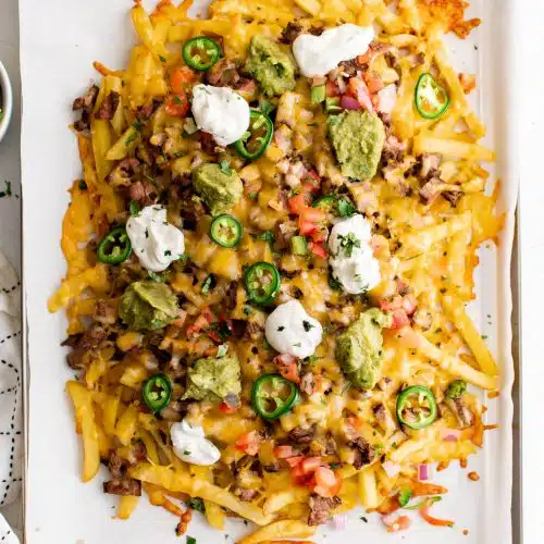 Large baking sheet line with parchment paper and filled with french fries smothered with carne asada, cheese, jalapeno, pico de gallo, sour cream, and guacamole.