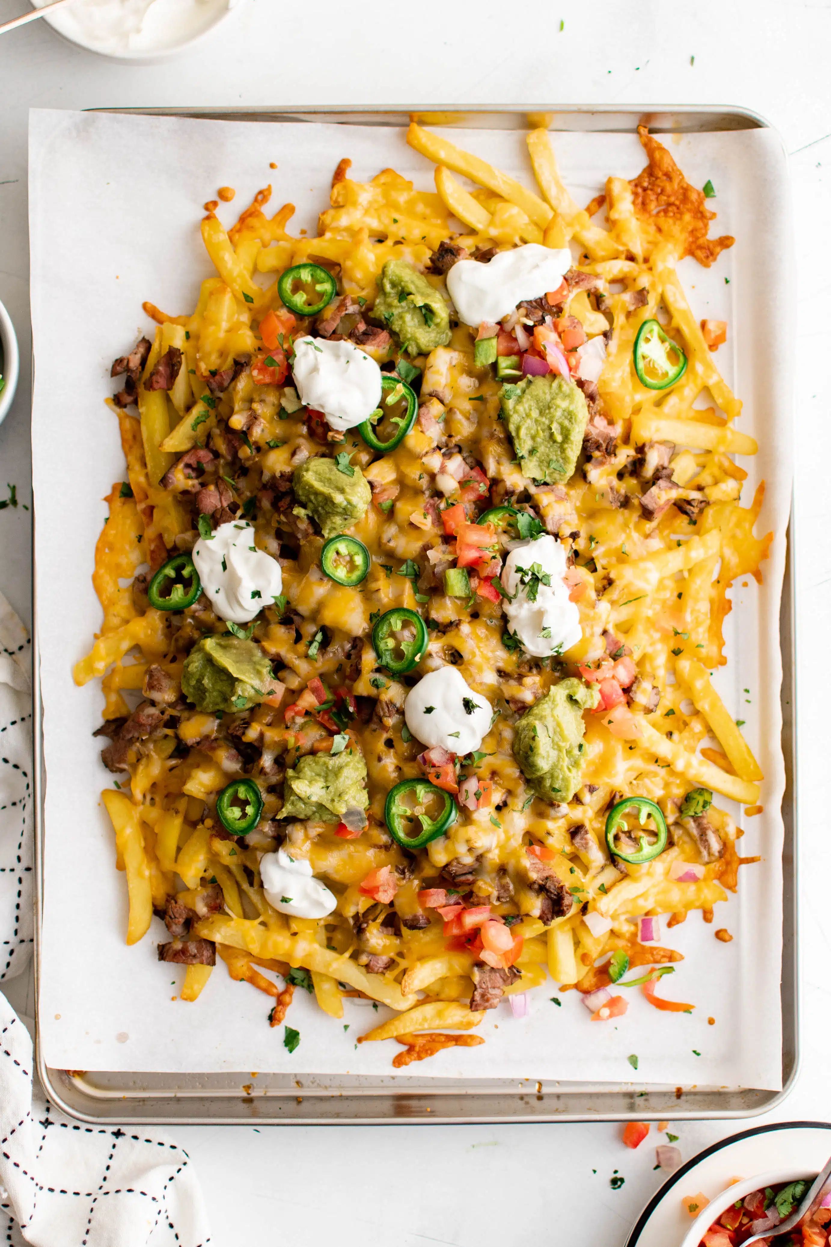Large baking sheet line with parchment paper and filled with french fries smothered with carne asada, cheese, jalapeno, pico de gallo, sour cream, and guacamole.