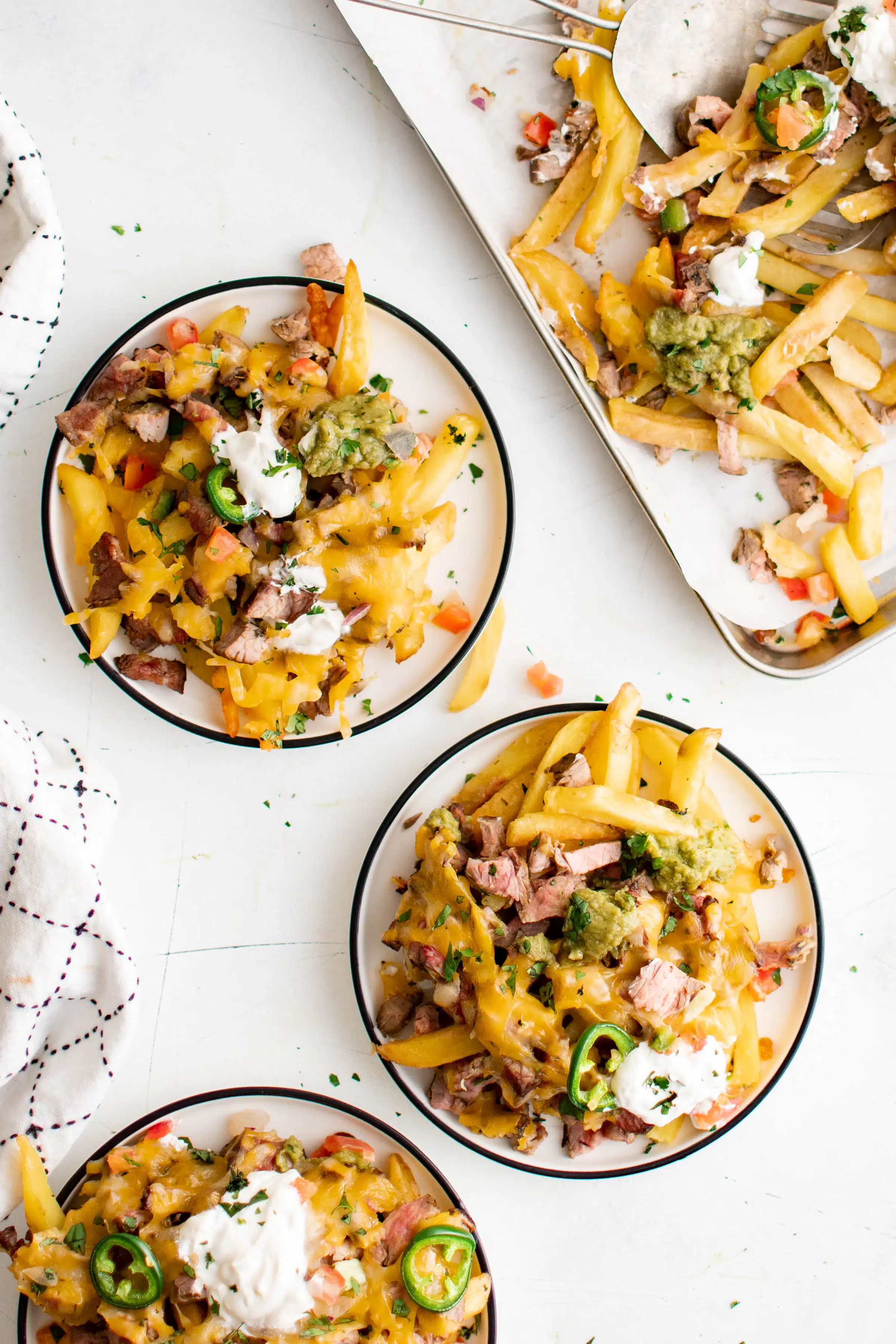 Three small serving plates filled with carne asada fries topped with sour cream next to a large baking sheet filled with the remaining carne asada nachos.