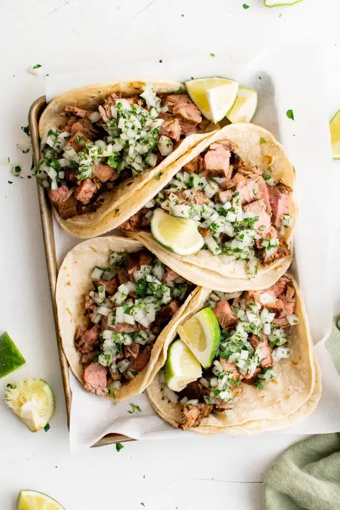 Tray filled with four carne asada tacos made with corn tortillas and topped with a fresh onion, cilantro, and lime juice salsa.