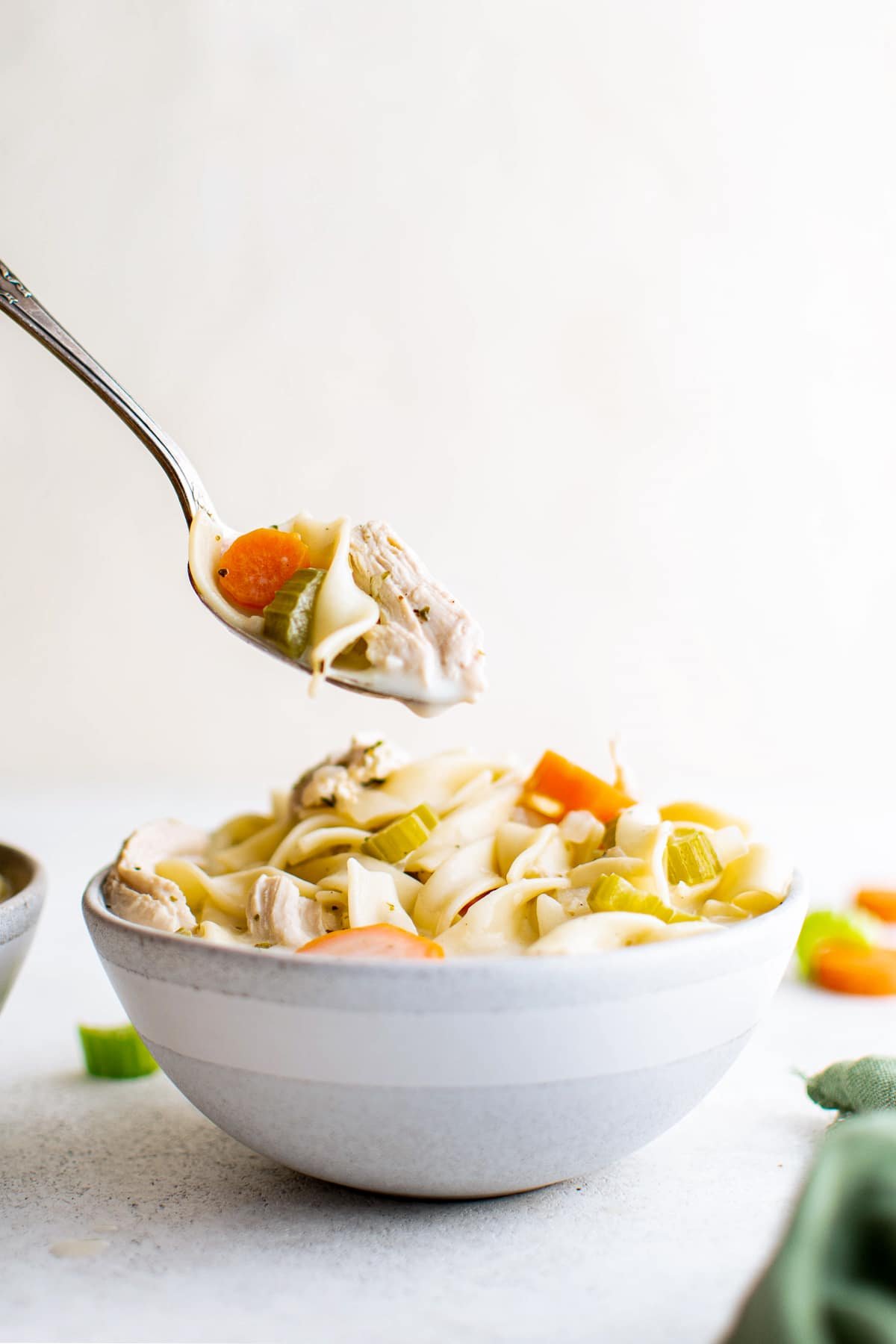 Metal spoon filled with creamy chicken noodle soup hovering above a white soup bowl filled with creamy chicken noodle soup.