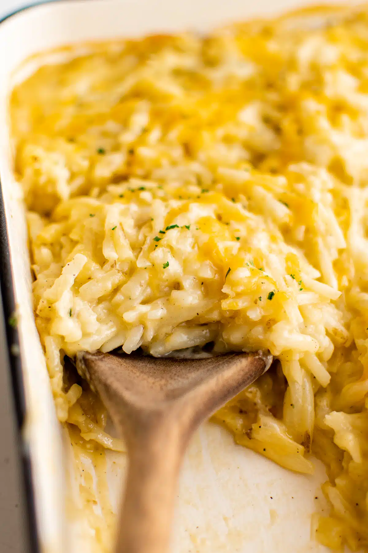 Wooden serving spoon scooping a large serving of cheesy hashbrown casserole from a white baking dish.