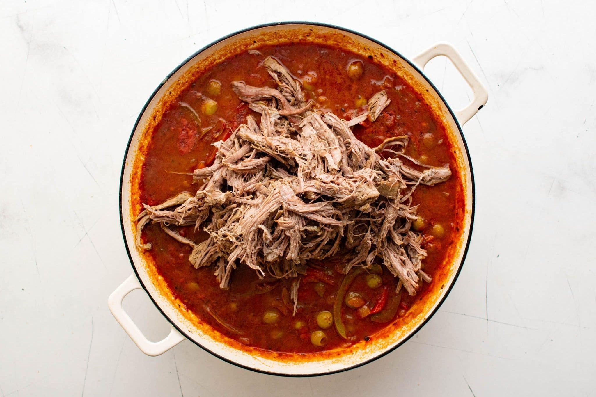Pile of shredded beef sitting on top of tomato and olive sauce in a Dutch oven.
