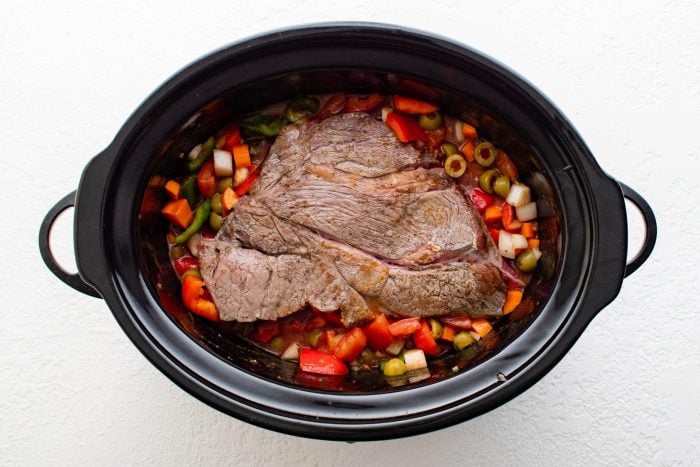 Seared chuck roast placed on top of a combination of vegetables, olives, broth, and seasoning in a large slow cooker.