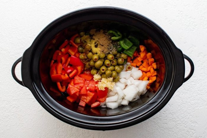 Large slow cooker filled with carrots, tomatoes, onion, olives, garlic, bell pepper, and seasoning.