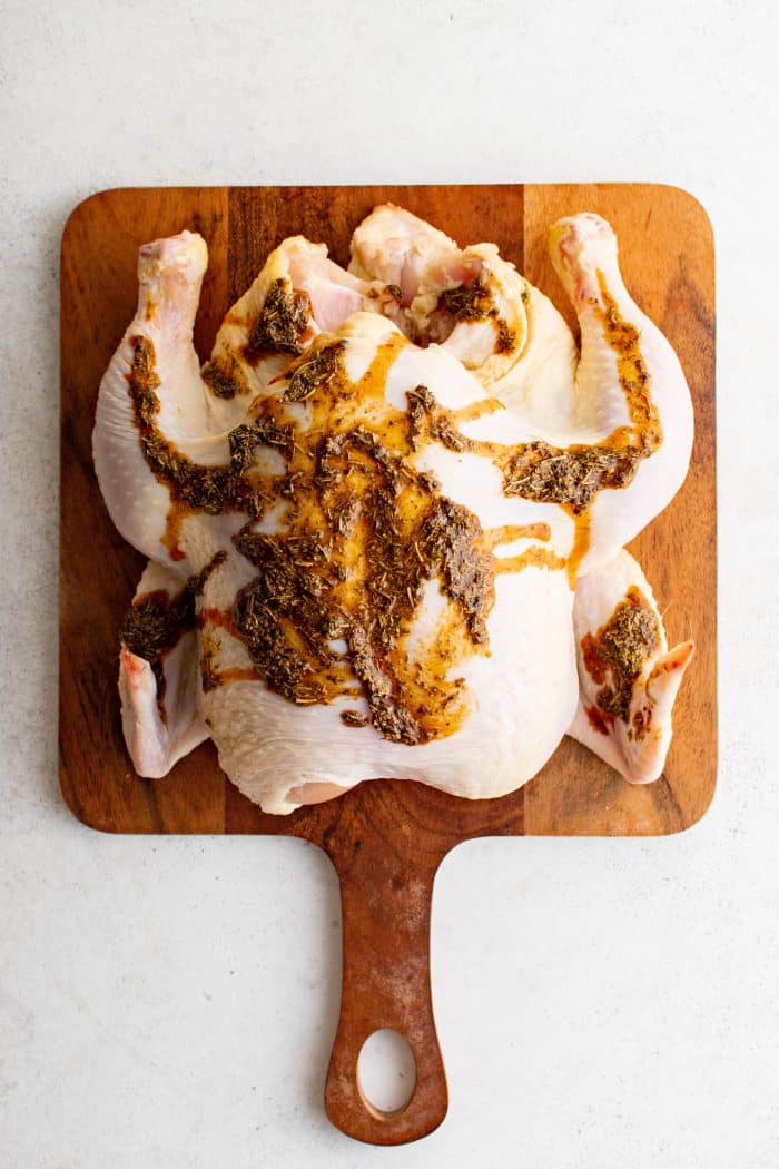 Whole chicken on a cutting board slathered with poultry seasoning in olive oil.