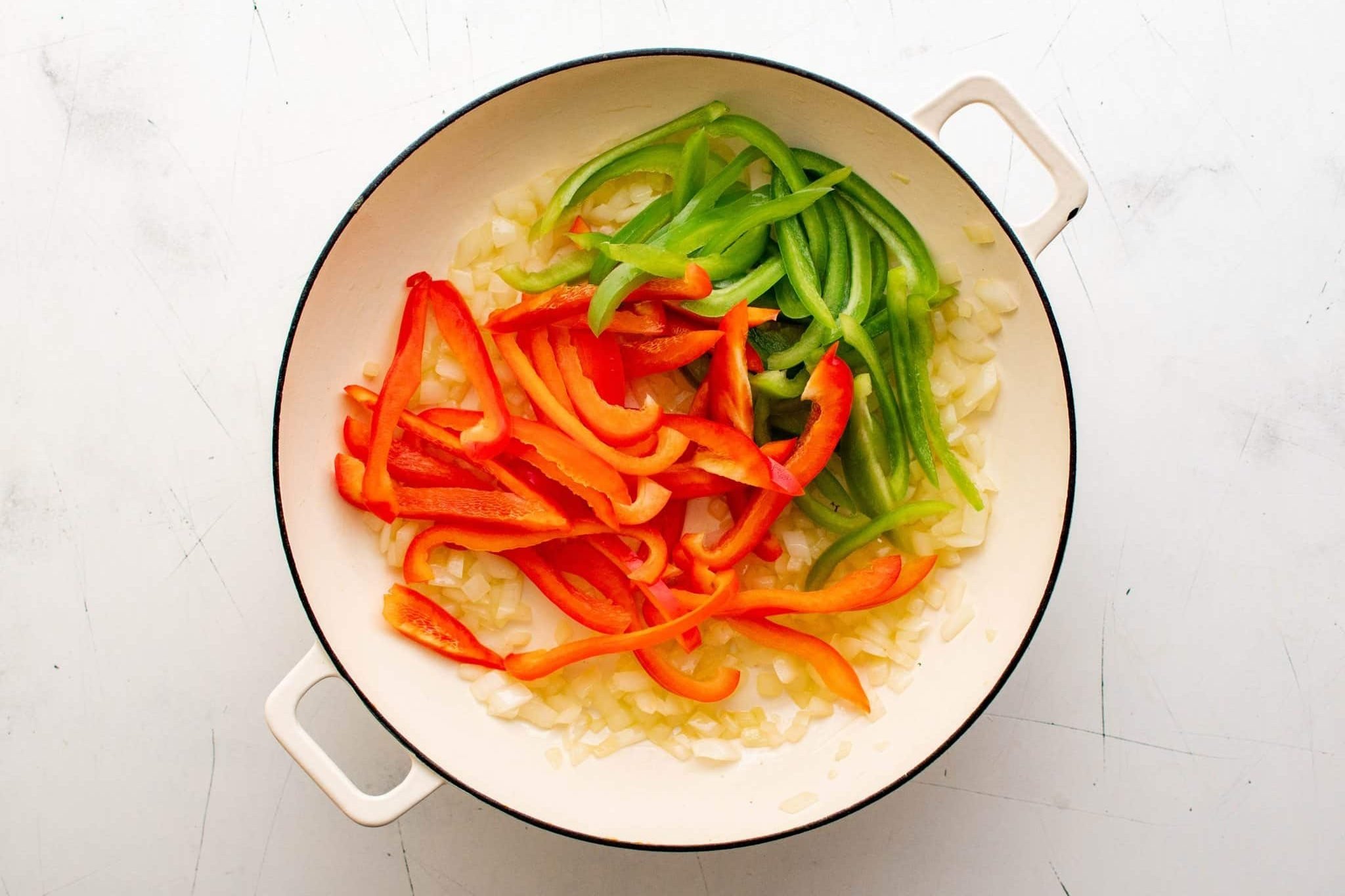 Sliced red and green bell peppers added to sautéed onions in a white skillet.