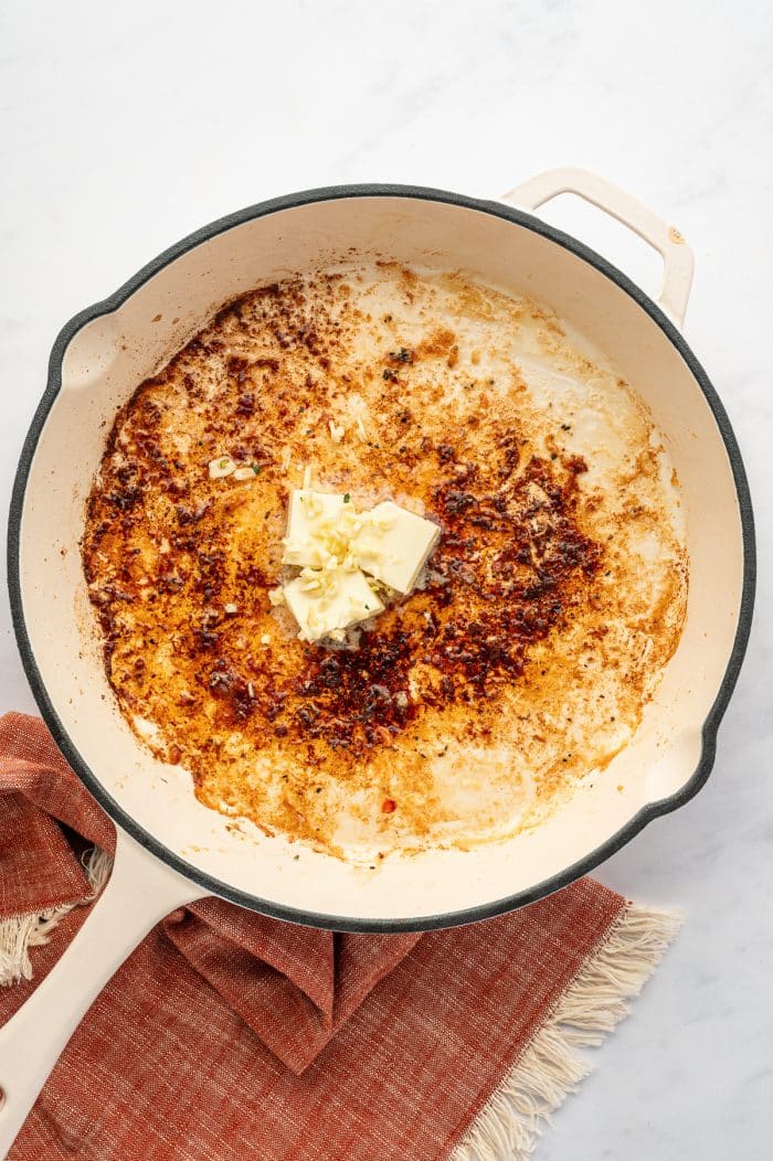 Overhead view of a cream-colored skillet with a black rim and light handle on a light gray marble surface. Two pats of butter and chopped garlic sit in the center of the pan over browned bits left from cooking the shrimp. A rust-orange fringed kitchen towel is tucked under the skillet.