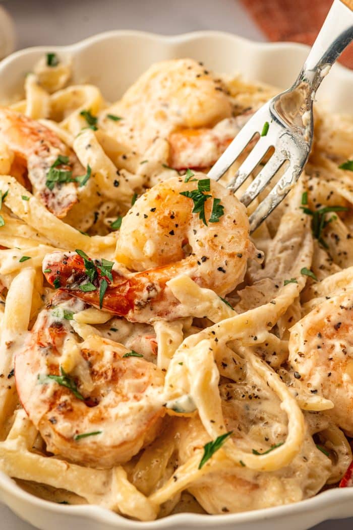 Close-up of creamy Shrimp Alfredo in a scalloped white bowl with a silver fork lifting a large shrimp from the pasta. The fettuccine is coated in Alfredo sauce and mixed with pink-orange shrimp, black pepper, and chopped fresh parsley. The background is softly blurred, with part of a rust-orange napkin visible in the upper corner.