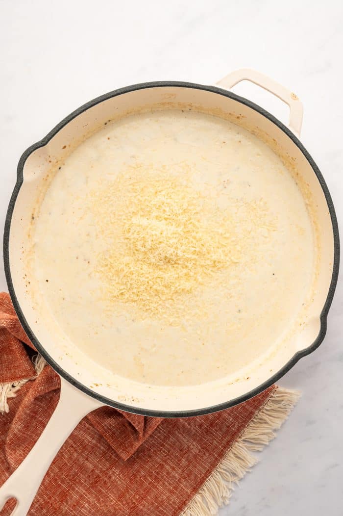 Overhead view of a cream-colored skillet with a black rim and light handle filled with creamy Alfredo sauce. A mound of finely grated Parmesan cheese sits in the center of the sauce. The skillet rests on a light gray marble surface with a rust-orange fringed kitchen towel underneath.