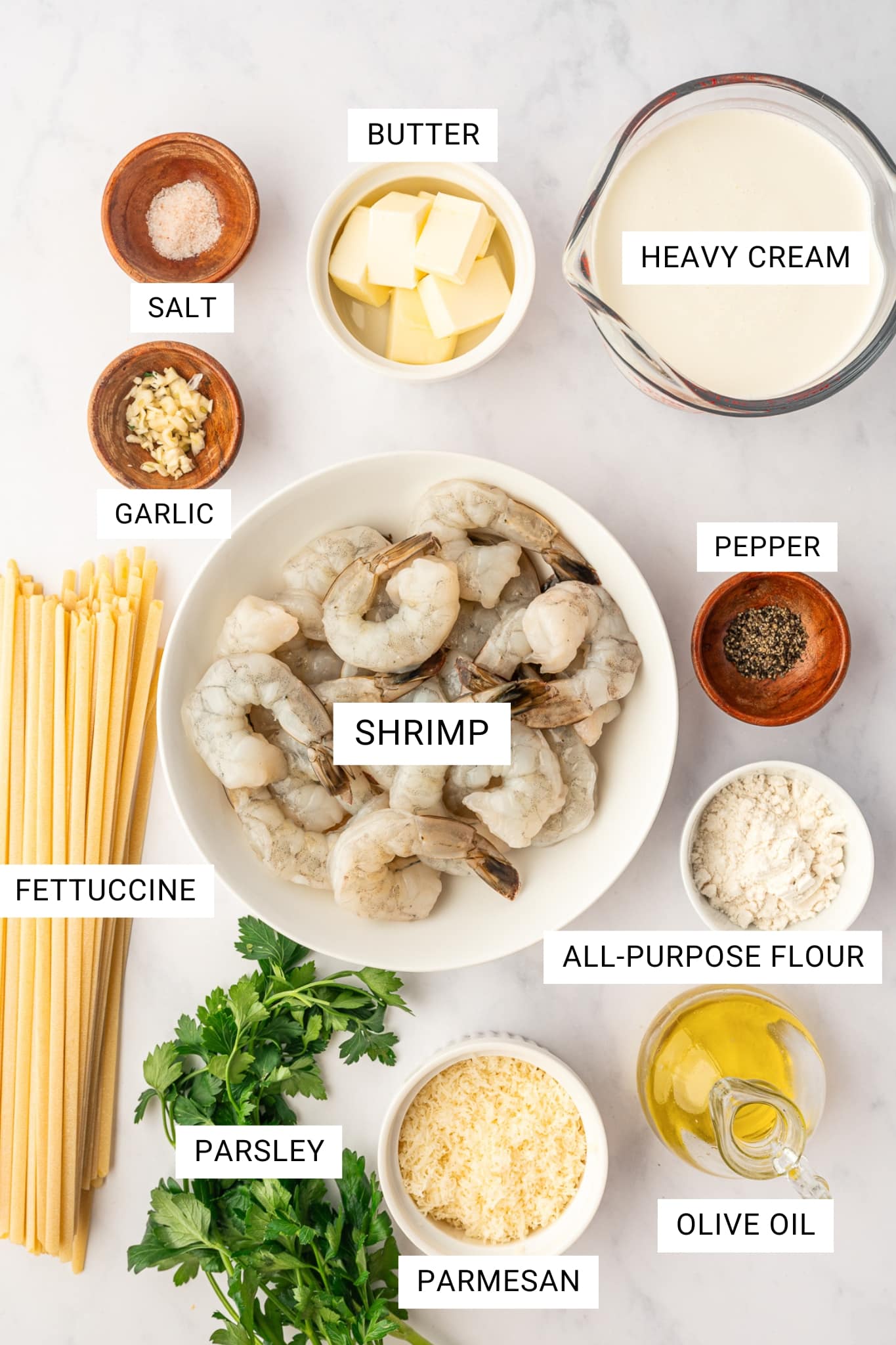 Overhead ingredient shot for Shrimp Alfredo on a light gray marble surface with labeled text overlays. A white bowl of raw shrimp sits in the center, surrounded by dry fettuccine, fresh parsley, grated Parmesan, olive oil in a glass bottle, all-purpose flour in a white ramekin, black pepper and salt in small wooden bowls, chopped garlic in a wooden bowl, butter in a white ramekin, and heavy cream in a glass measuring cup. Each ingredient is labeled with white text boxes reading FETTUCCINE, SHRIMP, PARSLEY, PARMESAN, OLIVE OIL, ALL-PURPOSE FLOUR, PEPPER, HEAVY CREAM, BUTTER, GARLIC, and SALT.