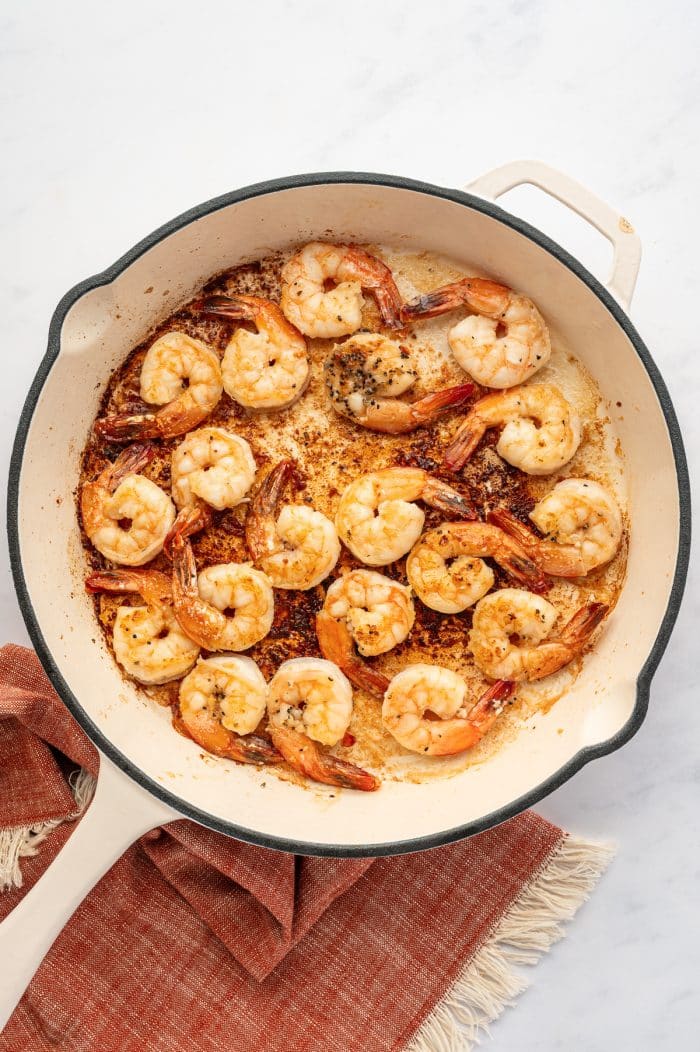 Overhead view of shrimp cooking in a cream-colored skillet with a black rim and light handle. The shrimp are pink and lightly golden, with several tails still attached. Browned bits and butter are visible on the bottom of the pan. A rust-orange fringed kitchen towel sits underneath the skillet on a light gray marble surface.