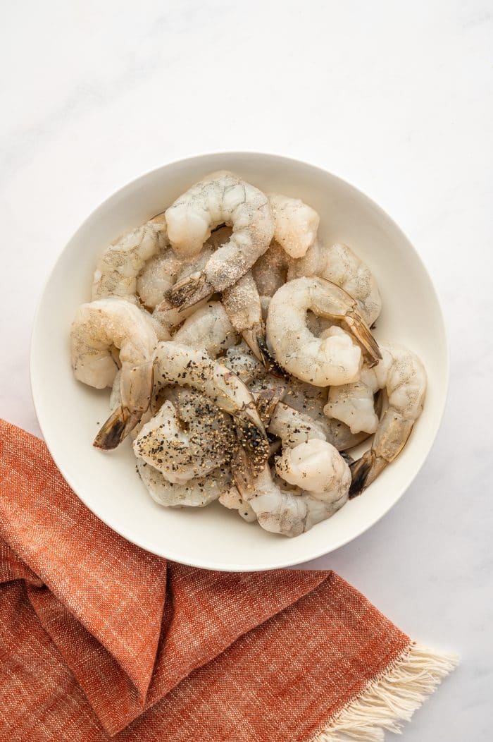 Overhead view of raw peeled shrimp in a round white bowl on a light gray marble surface. Some shrimp still have their tails on. The shrimp are lightly seasoned with black pepper and salt, and the bowl sits next to a rust-orange fringed kitchen towel.