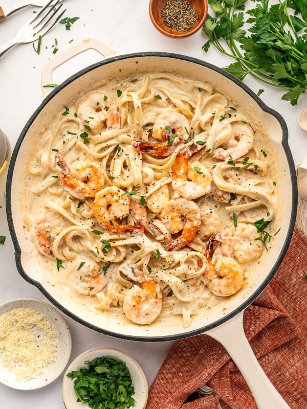 Overhead view of finished Shrimp Alfredo in a cream-colored skillet with a black rim and light handle. Fettuccine pasta is coated in creamy Alfredo sauce and topped with pink-orange shrimp, chopped fresh parsley, and black pepper. Surrounding the skillet are small bowls of grated Parmesan and chopped parsley, a wooden bowl of black pepper, fresh parsley sprigs, garlic cloves, two forks, and part of a glass at the edge of the frame. A rust-orange fringed kitchen towel sits under the skillet on a light gray marble surface.