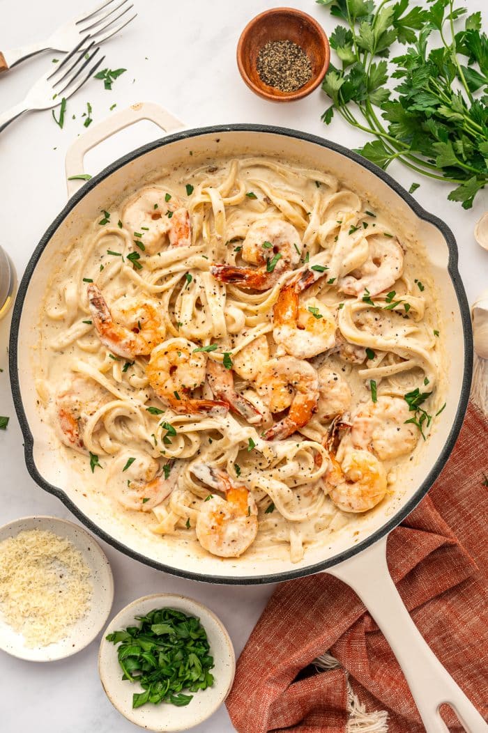 Overhead view of finished Shrimp Alfredo in a cream-colored skillet with a black rim and light handle. Fettuccine pasta is coated in creamy Alfredo sauce and topped with pink-orange shrimp, chopped fresh parsley, and black pepper. Surrounding the skillet are small bowls of grated Parmesan and chopped parsley, a wooden bowl of black pepper, fresh parsley sprigs, garlic cloves, two forks, and part of a glass at the edge of the frame. A rust-orange fringed kitchen towel sits under the skillet on a light gray marble surface.
