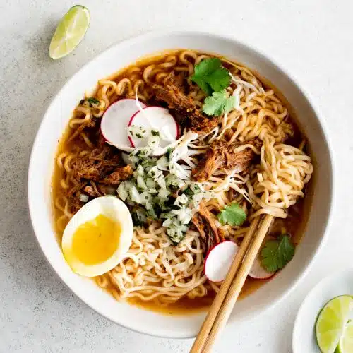 Large white serving bowl filled with birria ramen soup garnished with sliced radish, cilantro, soft boiled eggs, and salsa.