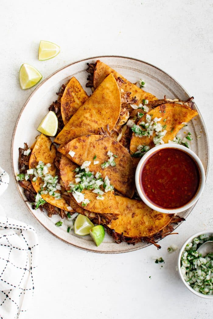 White plate filled with birria tacos garnished with onions ,cilantro, and lime wedges, and served with a small bowl of birria broth consumé.