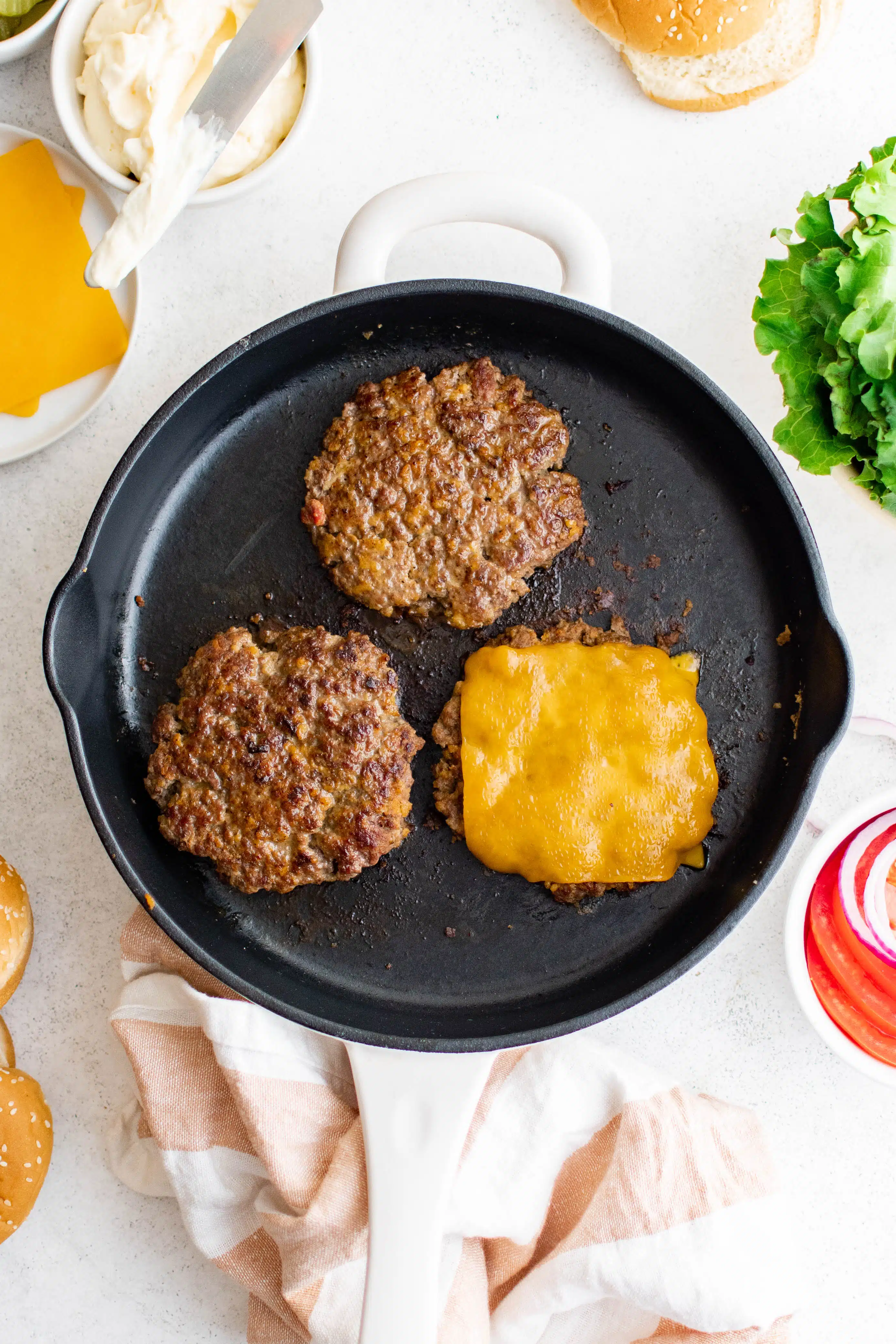Three hamburger patties (one with cheese) cooking in a large cast iron pan.