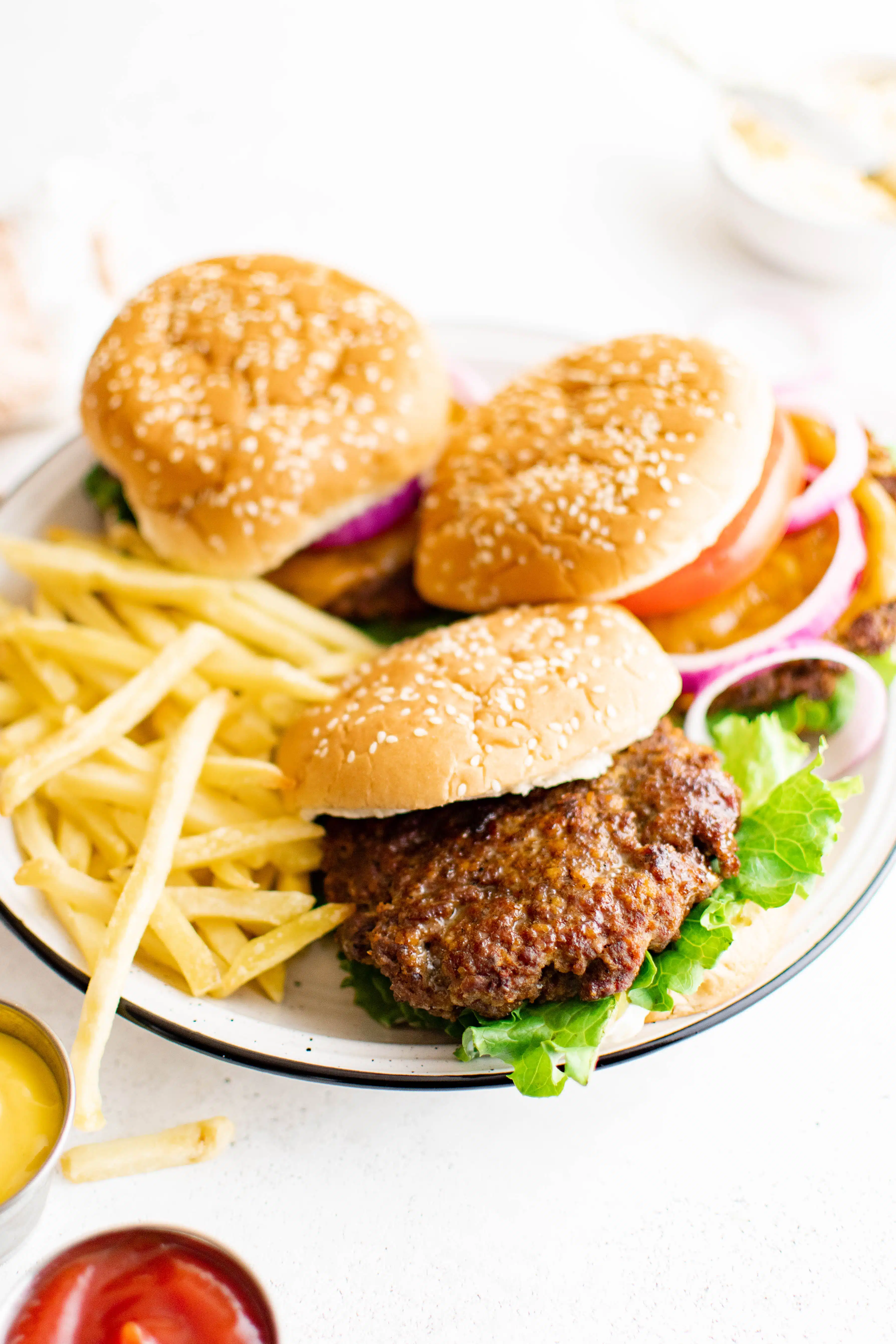 White plate with three prepared hamburgers on sesame buns with a side of potato fries.