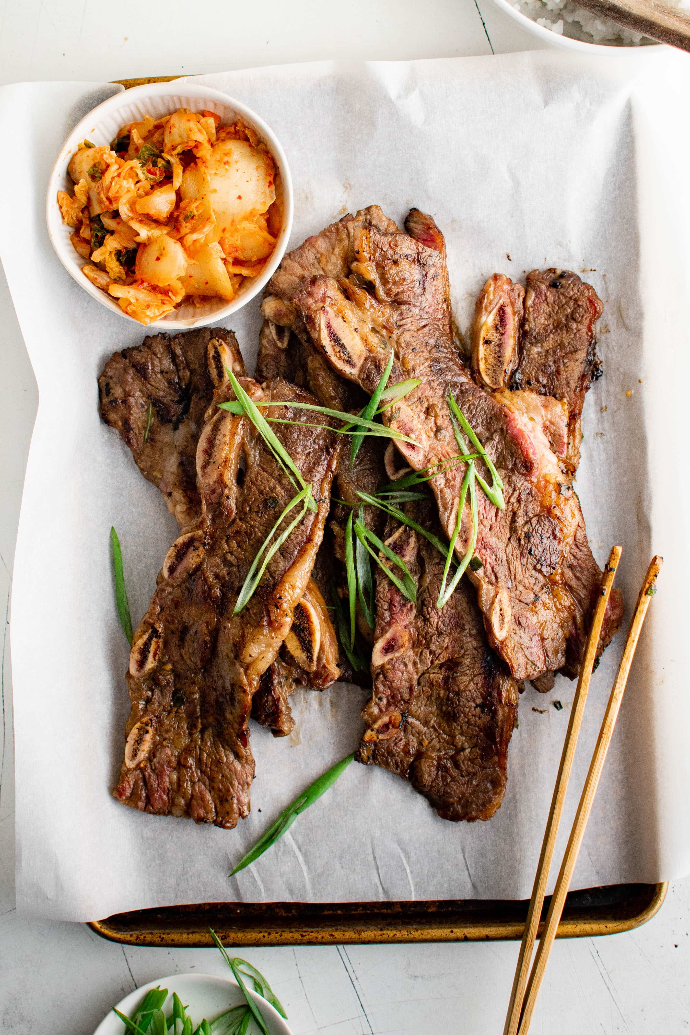 Grilled Kalbi (Korean BBQ Beef Short Ribs) on a baking sheet lined with parchment paper and garnished with thinly sliced green onions and served with a side of kimchi.
