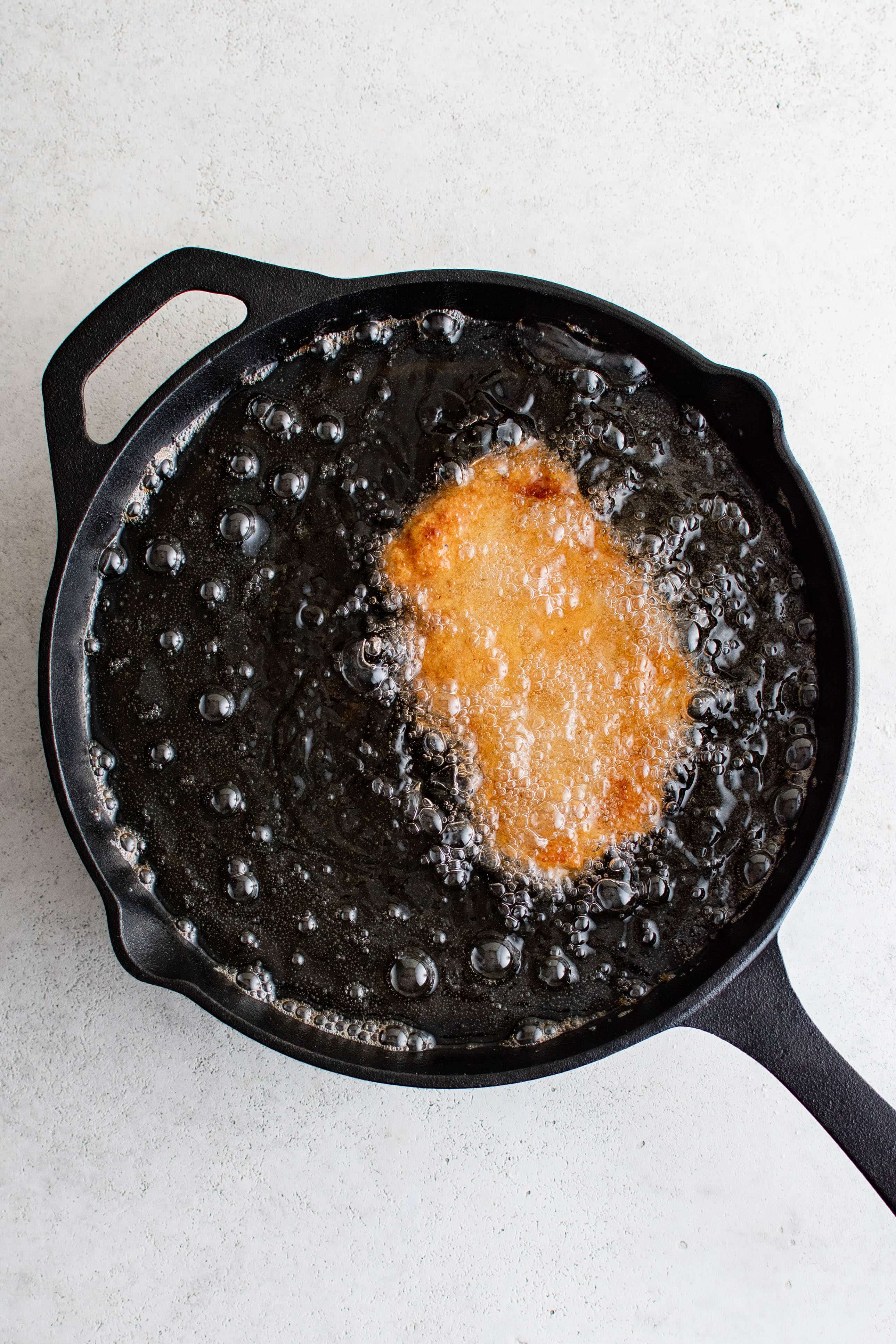 Breaded pork cutlet frying in a large cast iron skillet filled with hot oil.