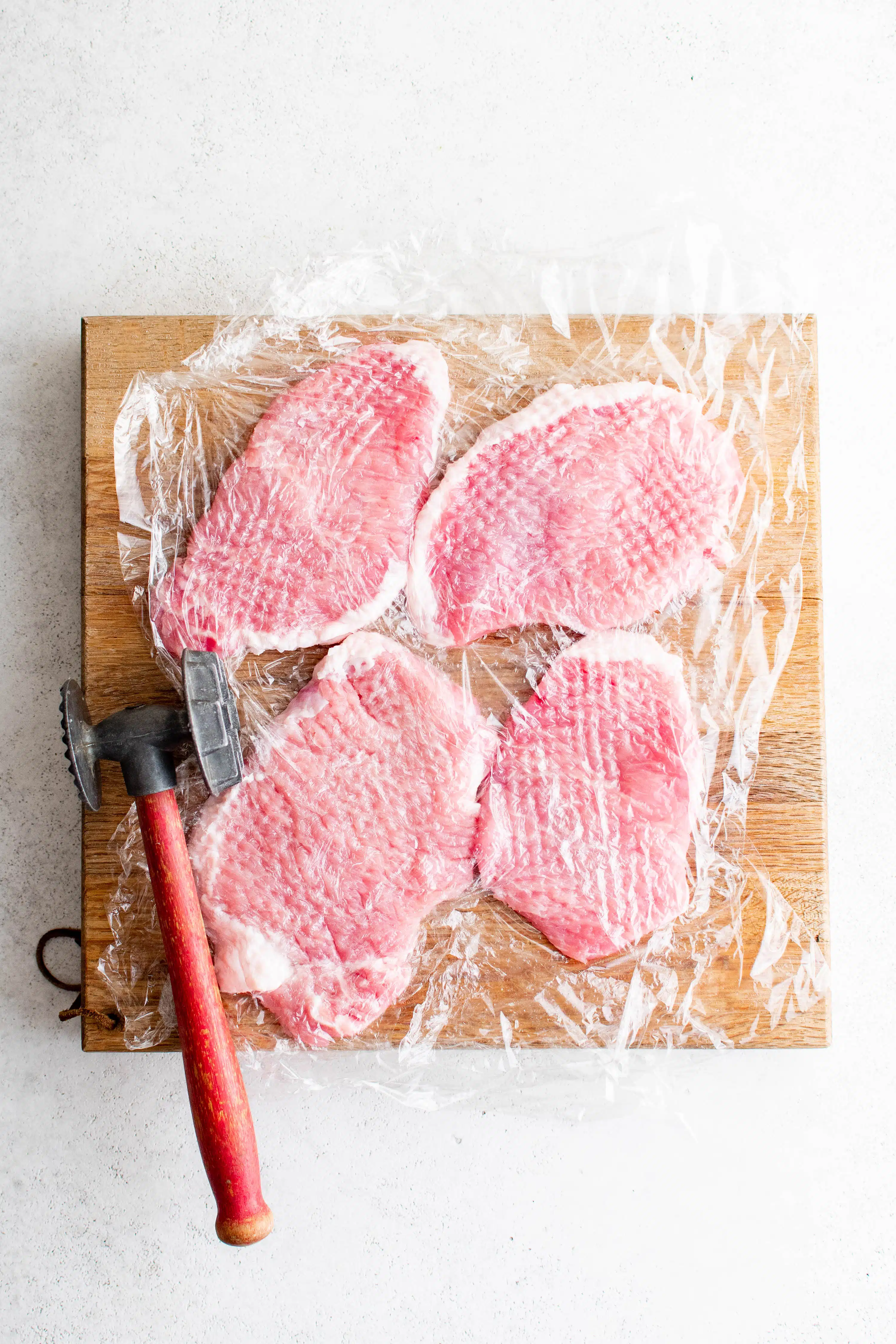 Four boneless pork chops on a wooden cutting board covered with plastic wrap with a meat tenderizer.