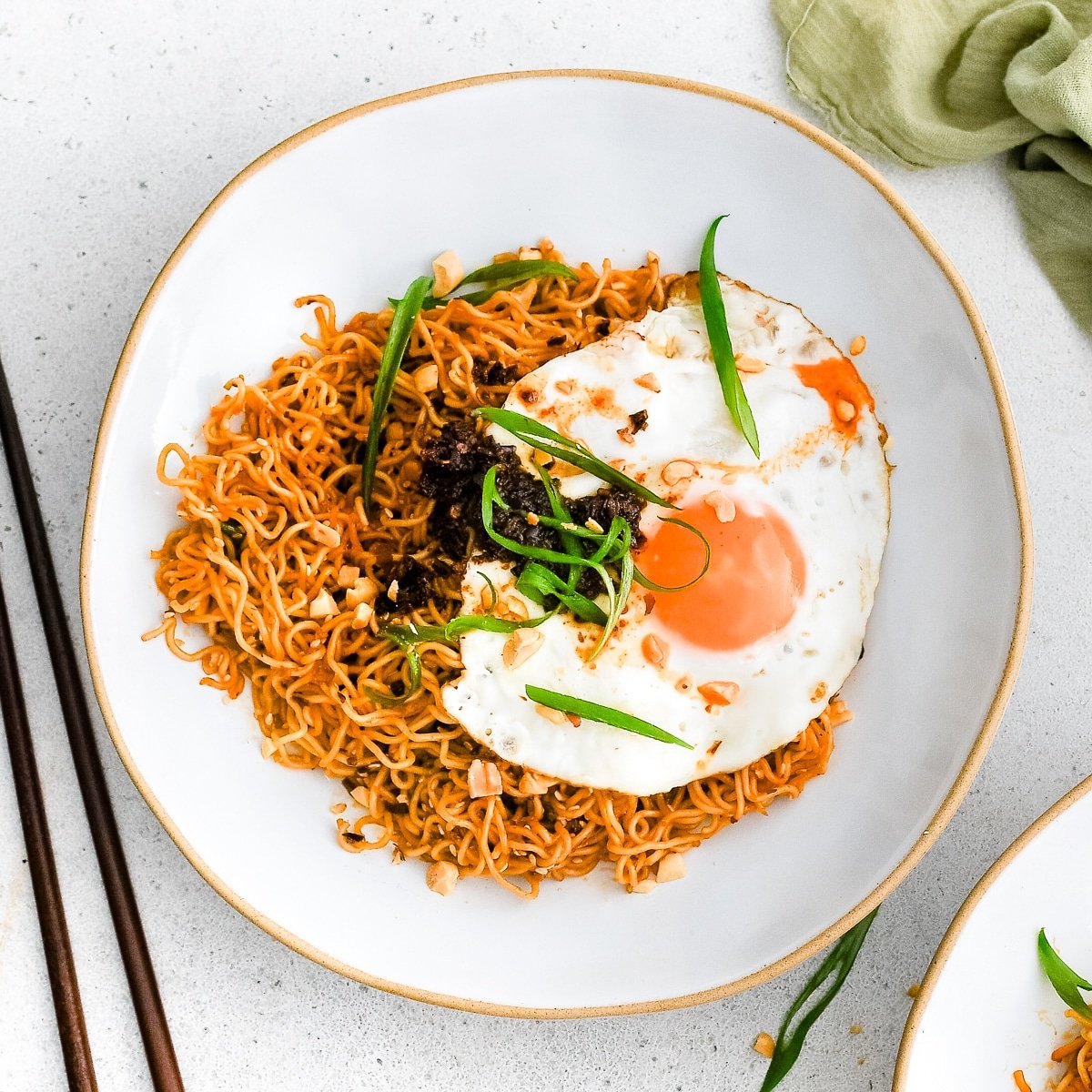 Shallow white serving bowl filled with spicy ramen noodles garnished with sliced green onions, chili crisp, and a beautiful fried egg.