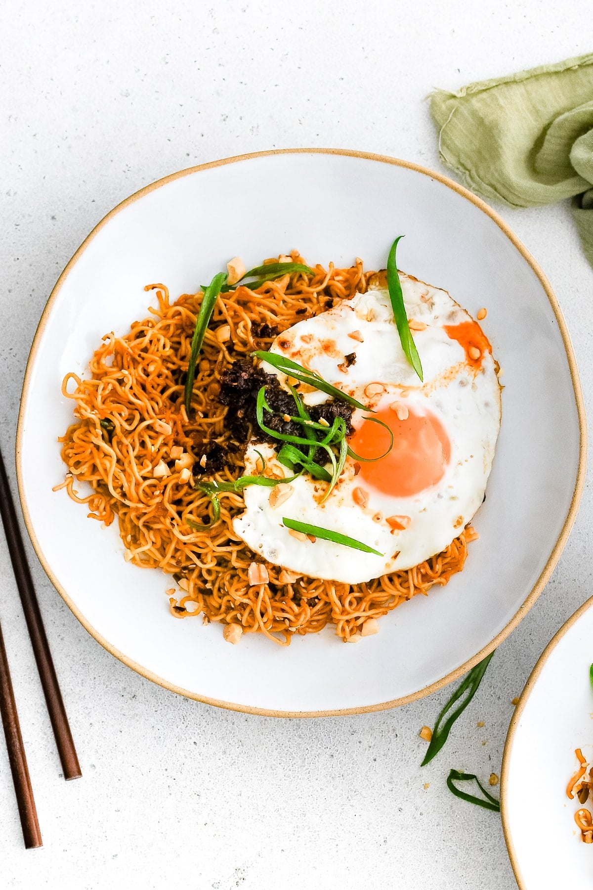 Shallow white serving bowl filled with spicy ramen noodles garnished with sliced green onions, peanuts, chili crisp, and a beautiful fried egg.