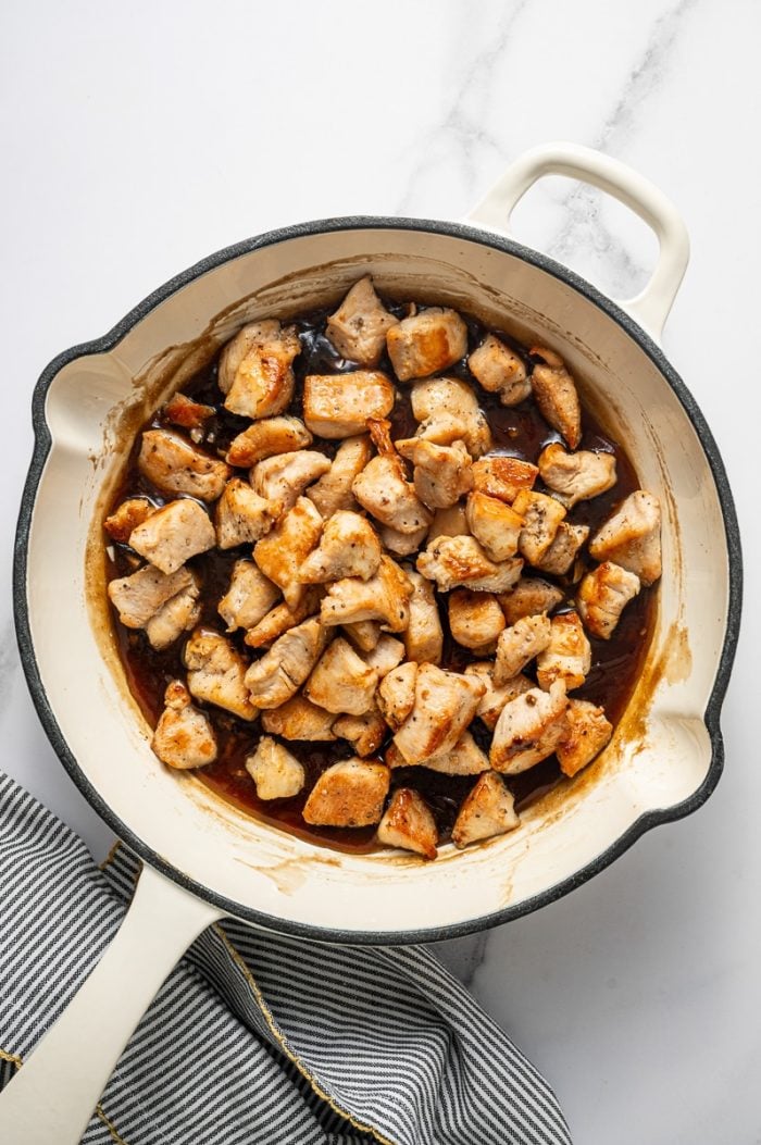 Overhead photo of browned chicken breast pieces added back into a cream-colored enameled skillet filled with dark teriyaki sauce, ready to be tossed, with a gray-and-white striped towel with yellow trim on the right and a white marble background.