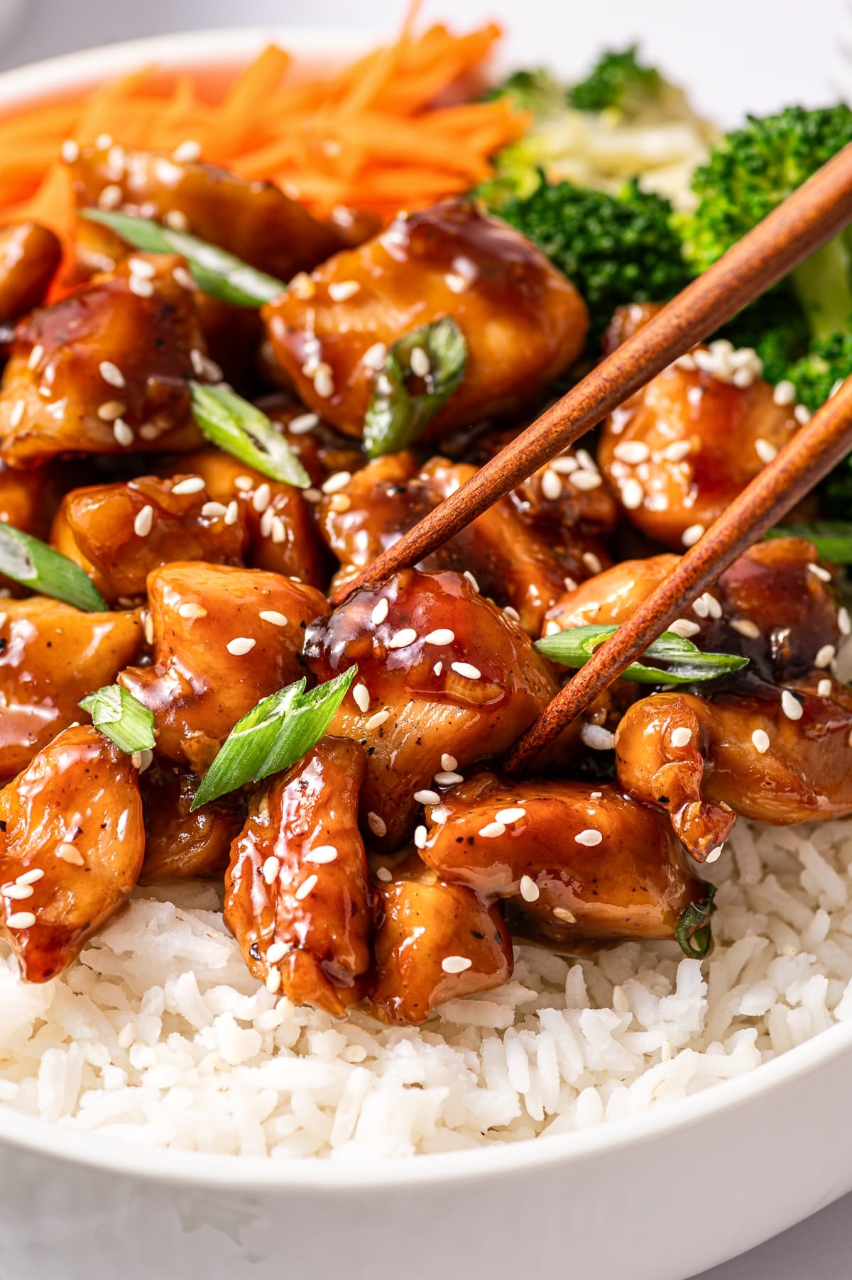 Close-up photo of glossy teriyaki chicken pieces on top of white rice, sprinkled with white sesame seeds and sliced green onions. A pair of wooden chopsticks is lifting a piece of saucy chicken, with julienned carrots and broccoli blurred in the background at the top of the bowl.