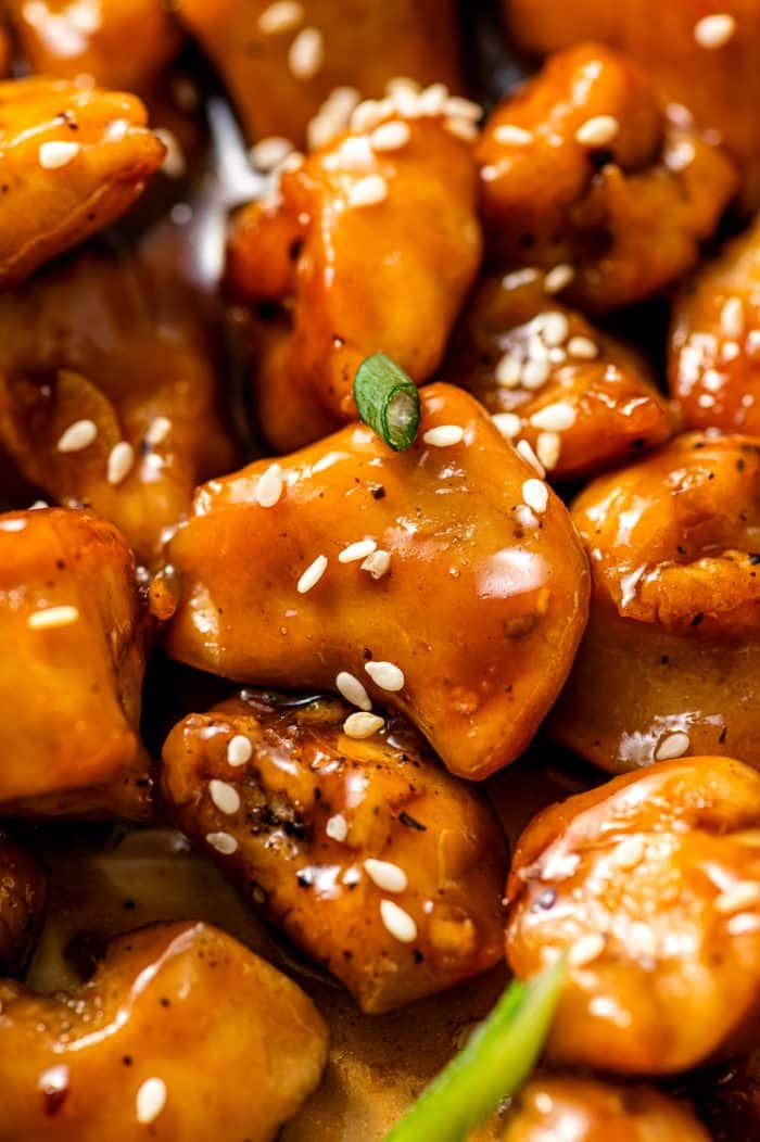 Extreme close-up of a single bite-size piece of teriyaki chicken coated in shiny brown glaze, topped with white sesame seeds and a small slice of green onion, with more glazed chicken pieces blurred in the background.