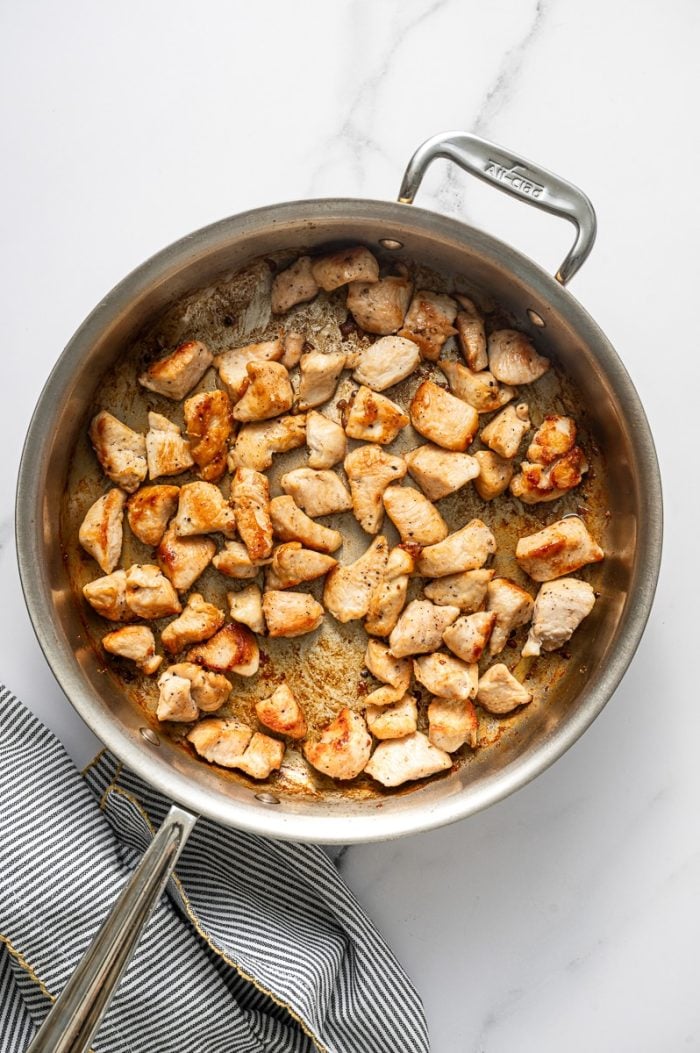 Overhead photo of bite-size chicken breast pieces searing in a stainless steel skillet with browned edges and caramelized bits on the pan, sitting on a white marble surface with a gray-and-white striped kitchen towel with yellow trim at the right edge.