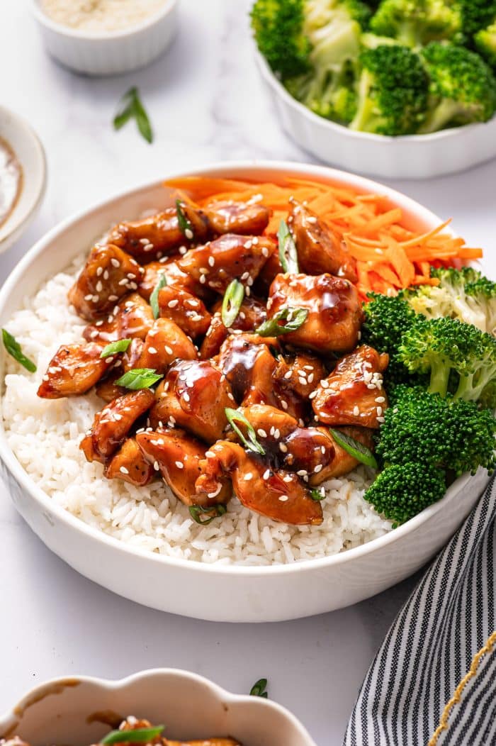 Side-angle photo of a white teriyaki chicken bowl with white rice topped with glossy teriyaki chicken, white sesame seeds, and sliced green onions, with a pile of julienned carrots and broccoli florets on the right side of the bowl; in the background are a white dish of broccoli and a small white ramekin on a white marble surface.