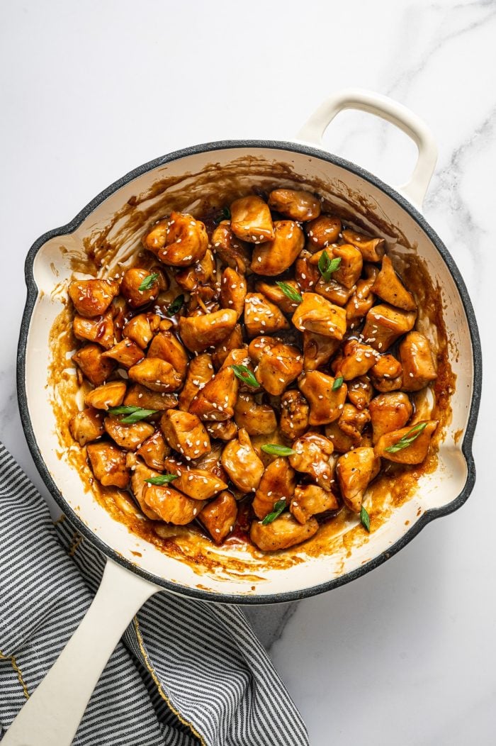 Overhead photo of teriyaki chicken bites coated in glossy brown sauce in a cream-colored enameled skillet, garnished with sliced green onions and white sesame seeds, on a white marble surface with a gray-and-white striped towel with yellow trim in the lower left.