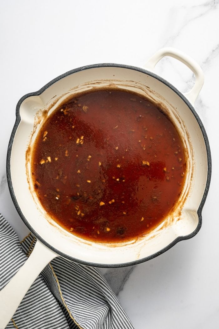 Overhead photo of a glossy brown teriyaki sauce simmering in a cream-colored enameled skillet with a black rim on a white marble surface, with small bits of garlic and ginger visible and a gray-and-white striped towel with yellow trim on the right.