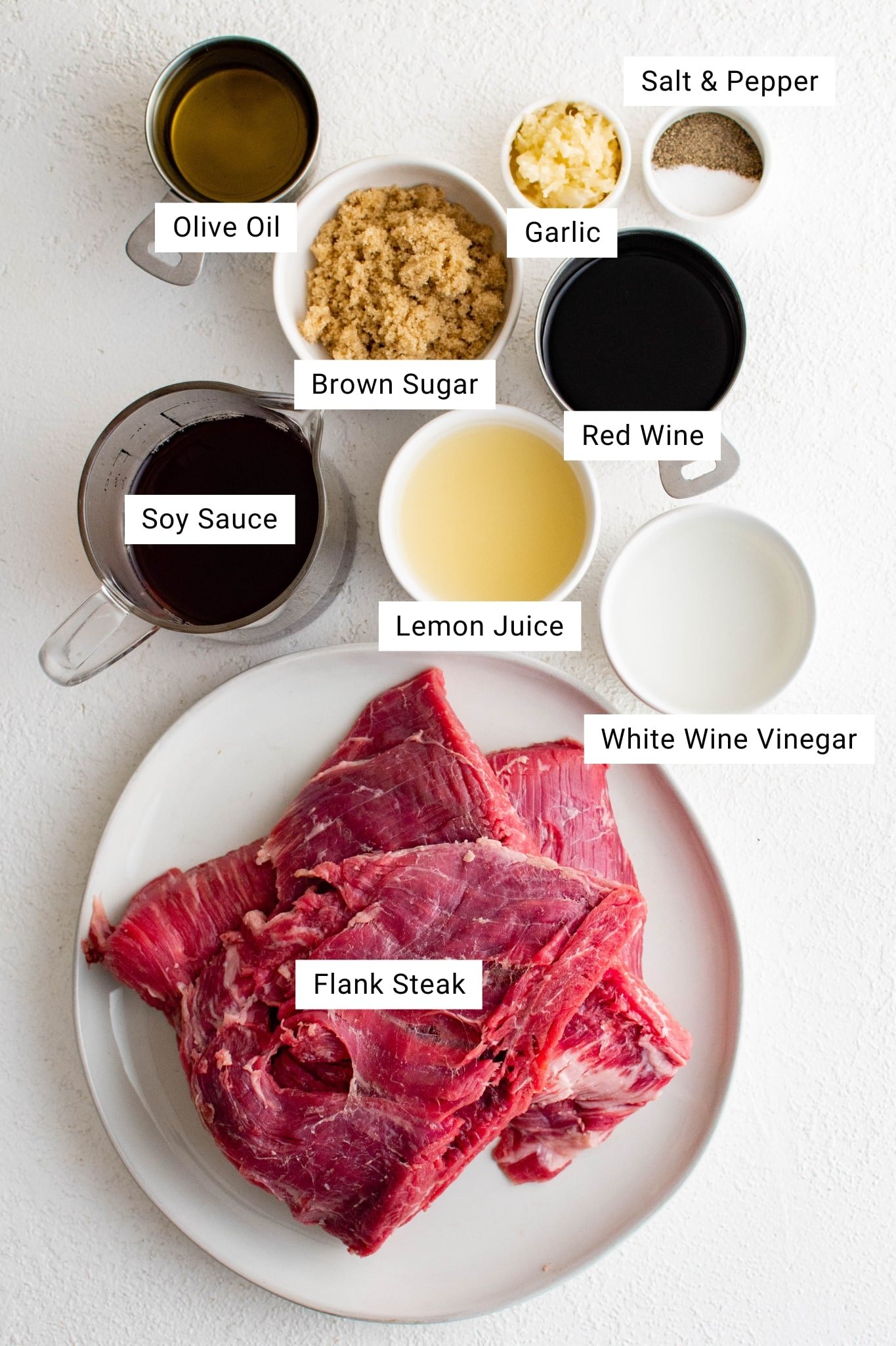 Overhead view of all the ingredients needed to make a homemade flank steak marinade arranged on a white surface. A round white plate at the bottom holds raw flank steak with visible grain and muscle fibers. Around the plate are small bowls and measuring cups labeled with their contents: olive oil in a metal measuring cup, light brown sugar in a white bowl, minced garlic in a small dish, red wine in a glass measuring cup, soy sauce in a dark liquid-filled measuring cup, lemon juice in a white bowl, white wine vinegar in another small white bowl, and a tiny dish of salt and ground black pepper. Each ingredient is clearly labeled with text overlays.