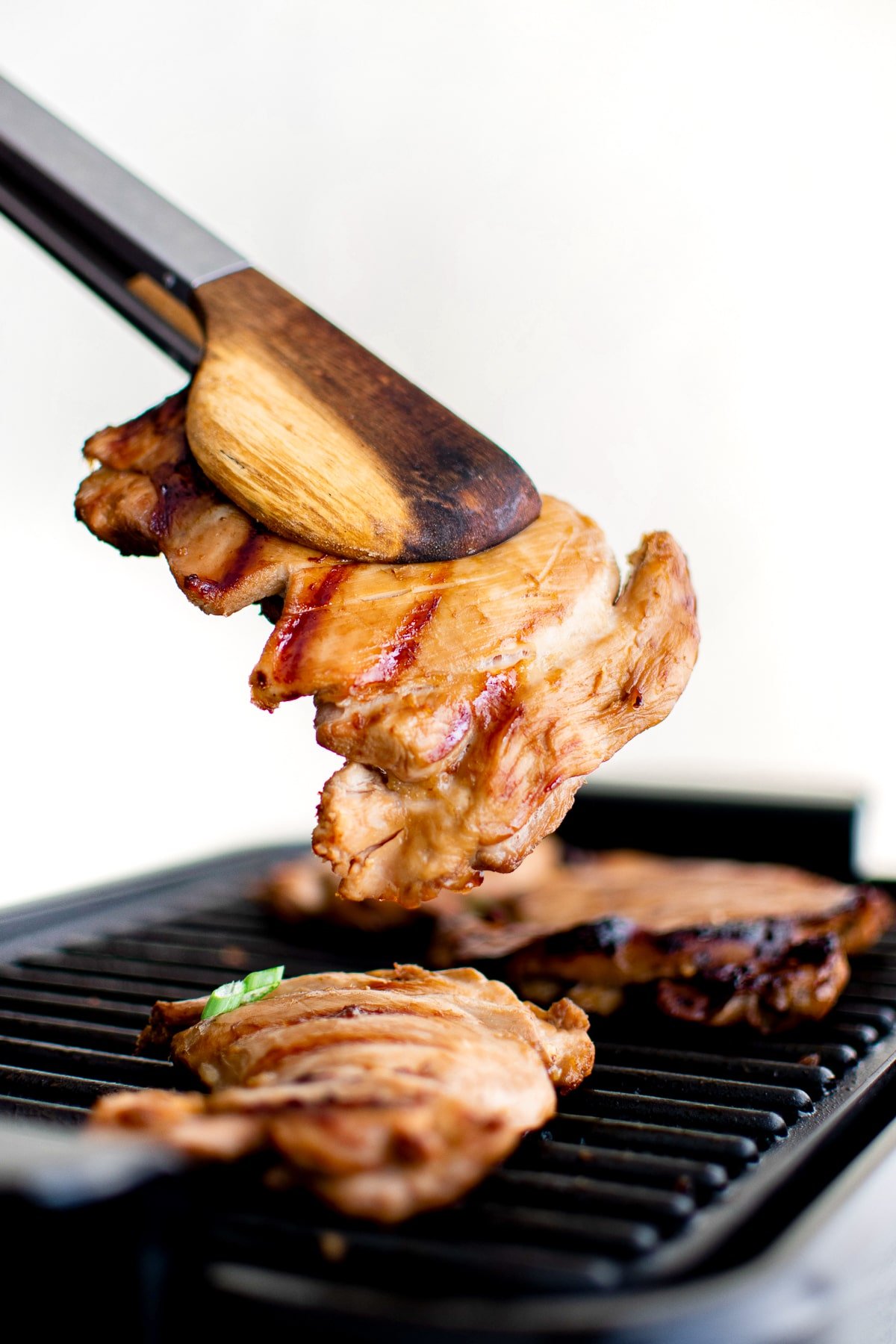 Wooden tongs lifting a piece of grilled Hawaiian chicken from a grill pan.