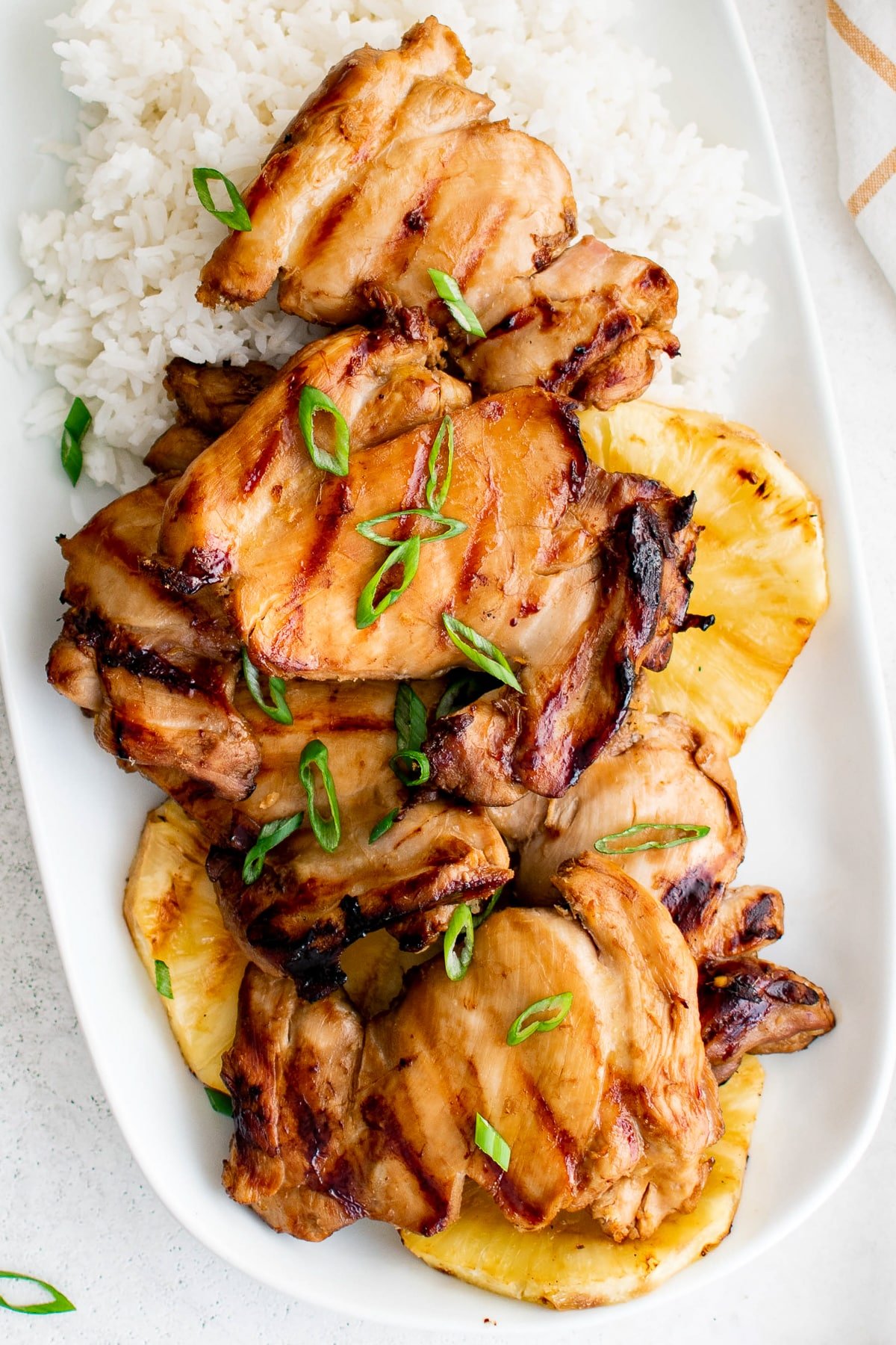 A platter of grilled Hawaiian chicken thighs served with white rice, grilled pineapple slices, and green onions.