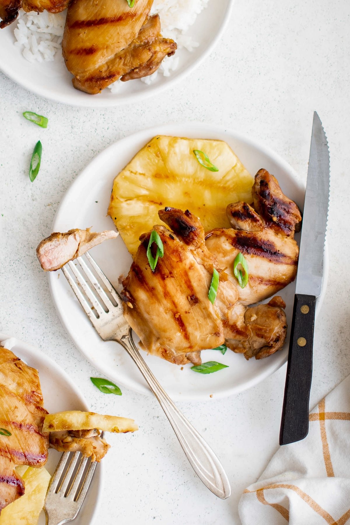A dinner plate with grilled Hawaiian chicken, white rice, and a slice of grilled pineapple, served with fork and knife.