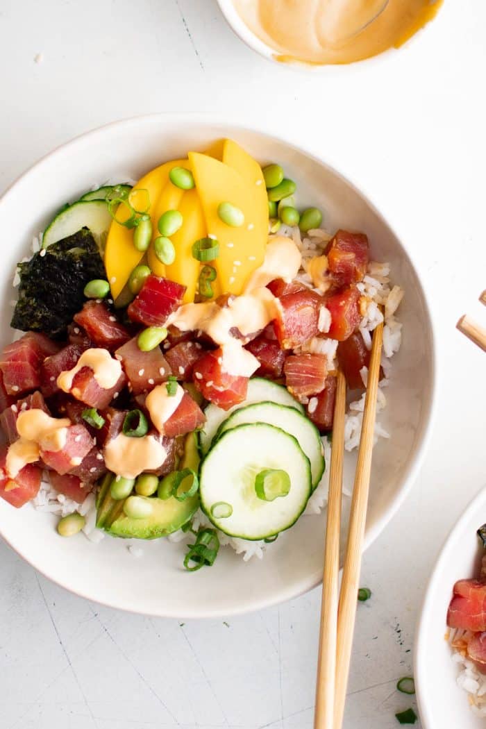A vibrant poke bowl featuring sushi-grade ahi tuna, creamy avocado slices, sweet mango chunks, edamame, crisp cucumber, and green onions, all drizzled with spicy Sriracha mayo atop a bed of sticky white rice.