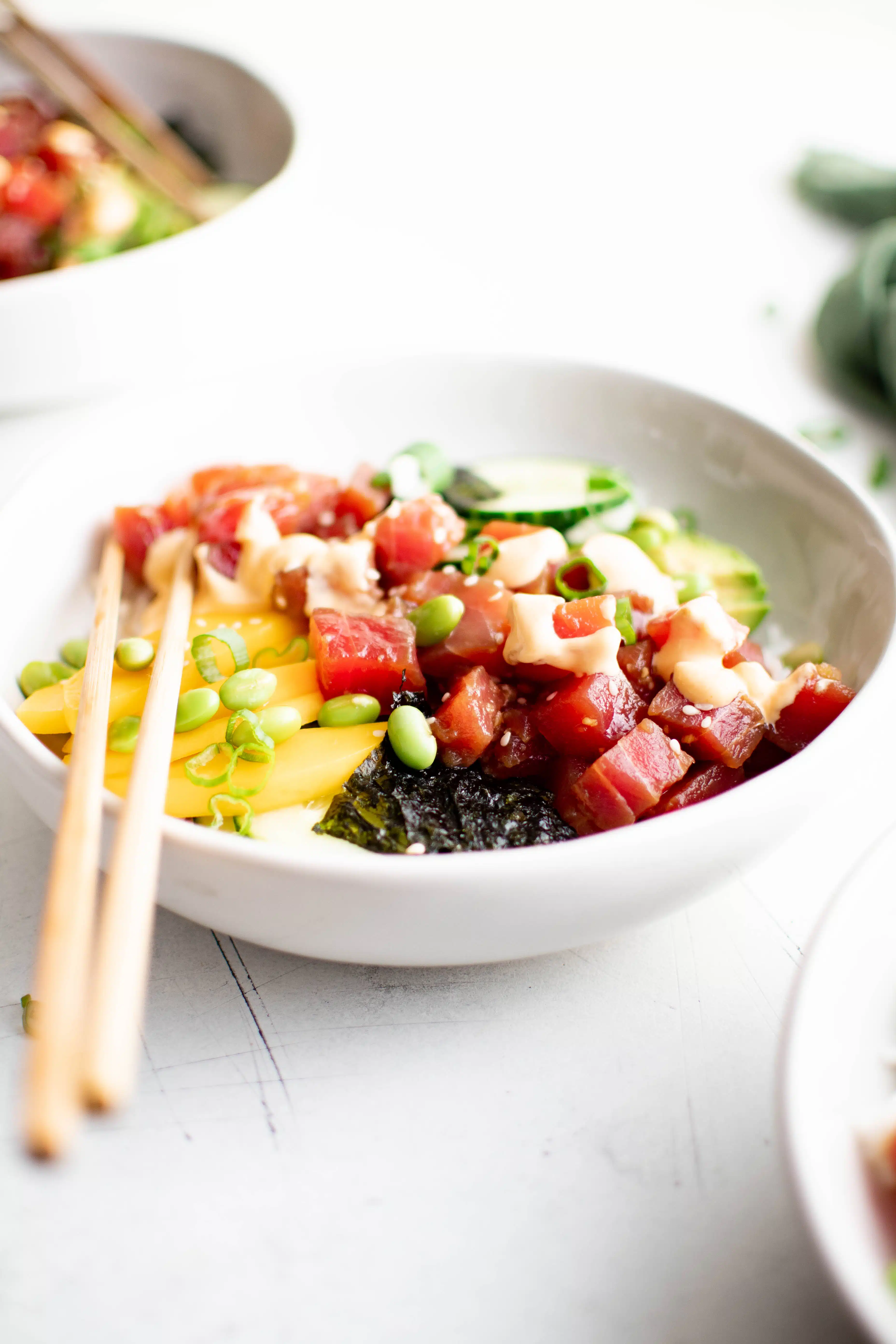 White bowl filled with white rice topped with sliced cucumber, avocado, mango, ahi tuna, green onions, edamame, and drizzled with creamy Sriracha mayo sauce.