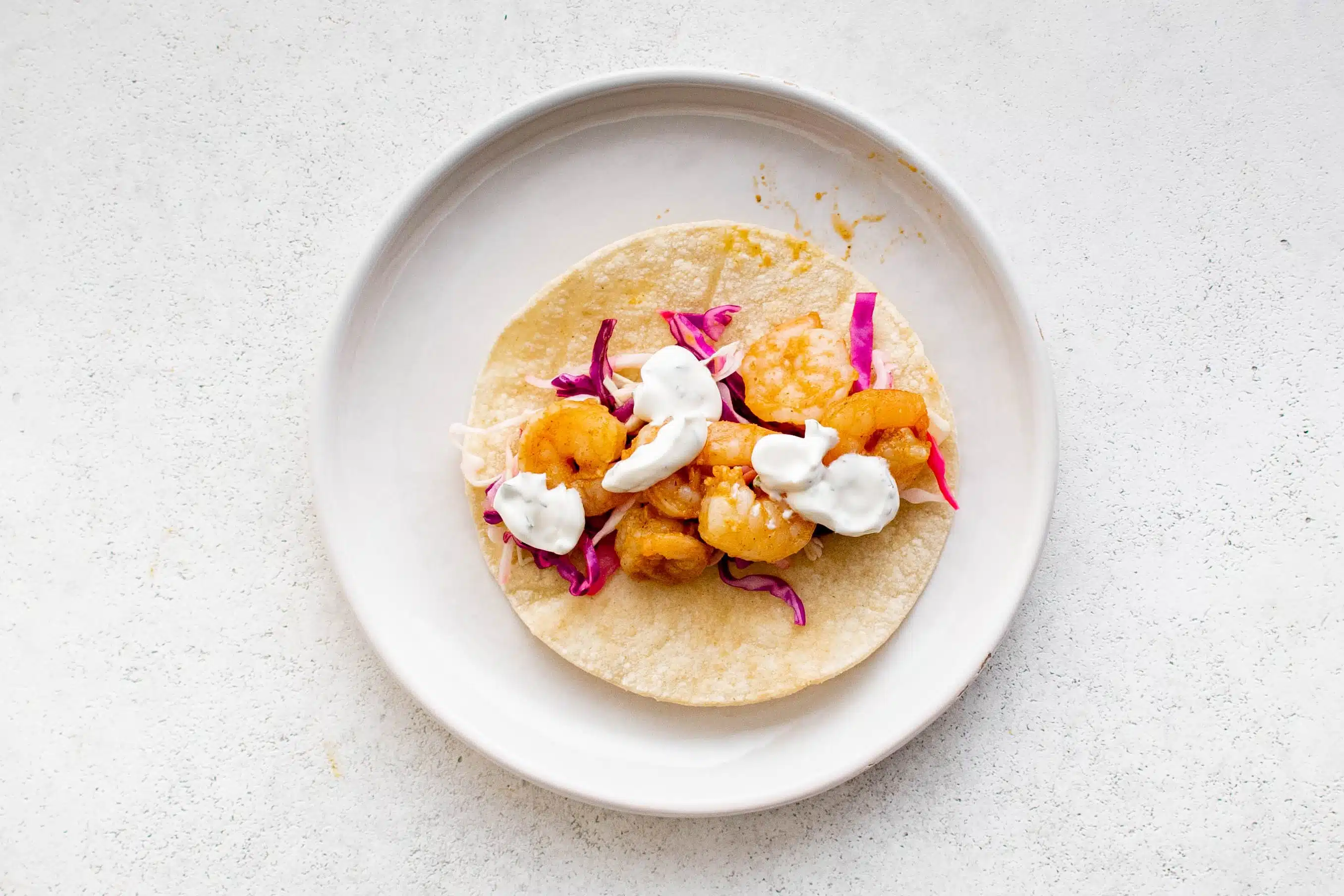 Tortilla on a white plate topped with a light cabbage slow, cooked shrimp, and creamy shrimp taco sauce.
