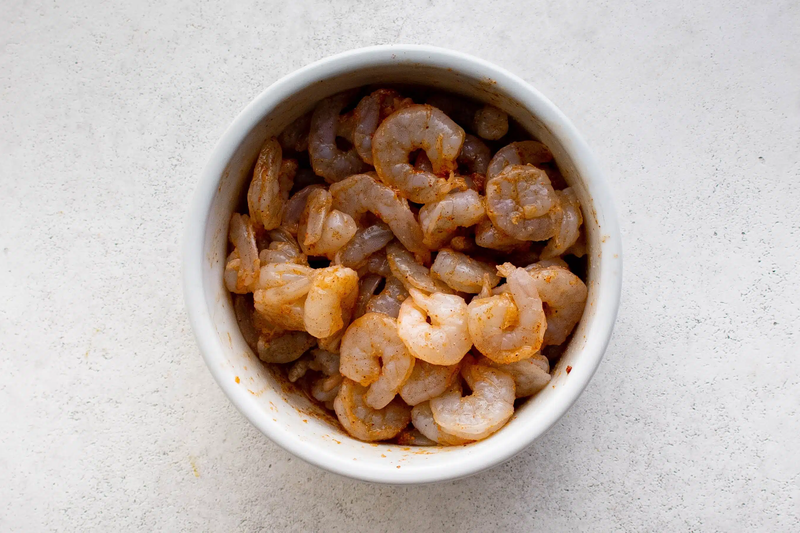 White bowl filled with raw shrimp seasoned with salt, chili powder, paprika, onion powder, garlic powder, and olive oil.